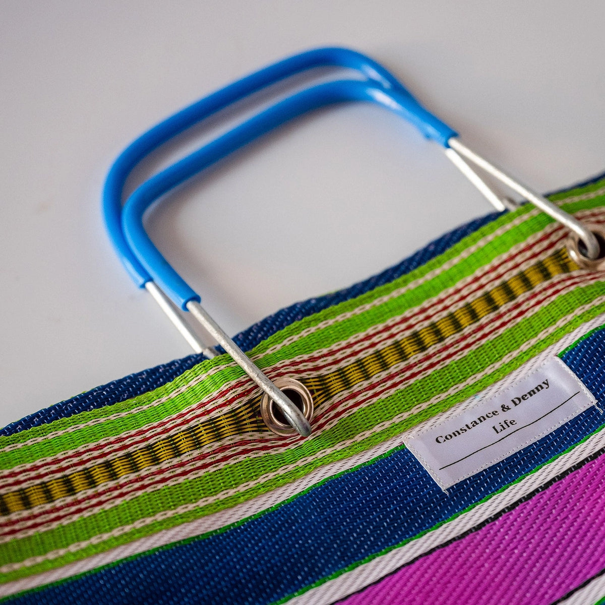 Close-up of a colorful striped fabric tote bag with blue metal handles and a white label reading 'Constance & Denny Life'.