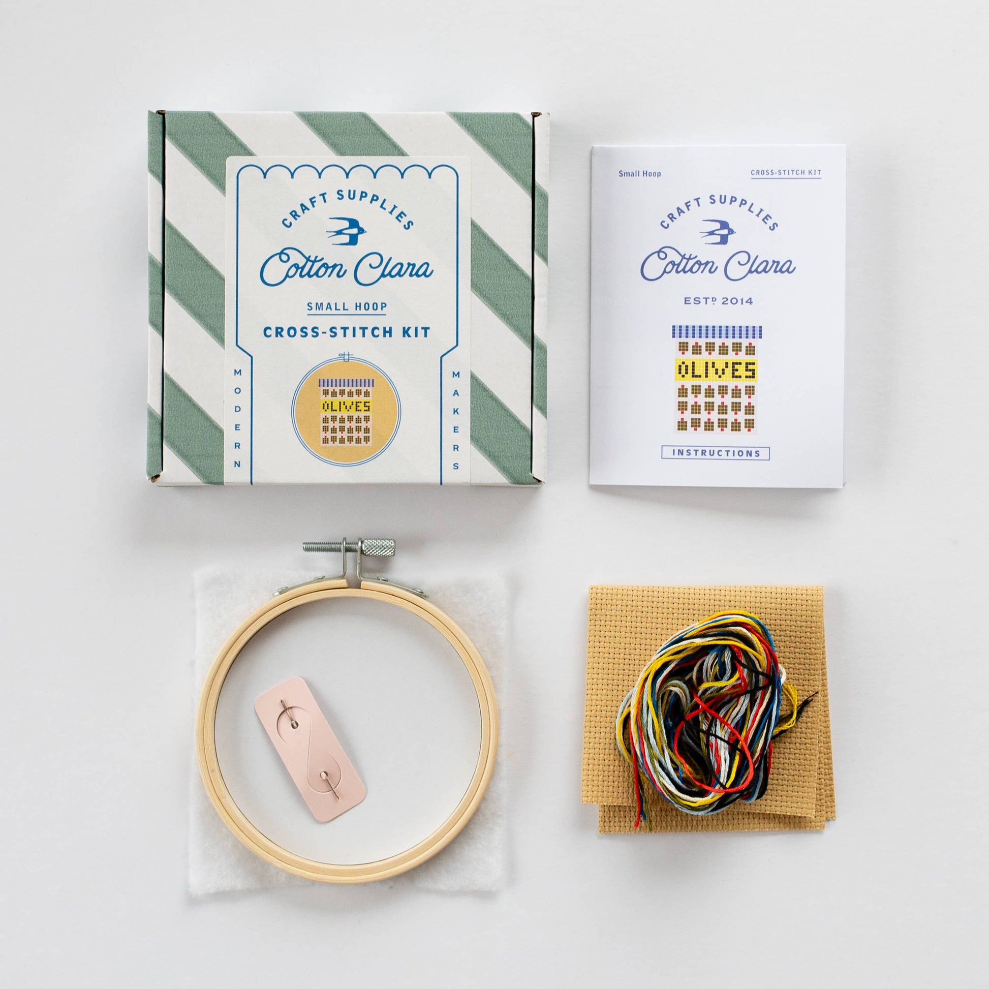 Contents of the Olives Mini Cross Stitch Kit laid out: coloured Aida, embroidery threads, needle, instructions, wadding, and backing disc.