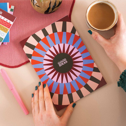 Colourful notebook with geometric design and 'Good Ideas' text, held by a person with a cup of coffee on a pink surface. UK Made. Eco friendly.