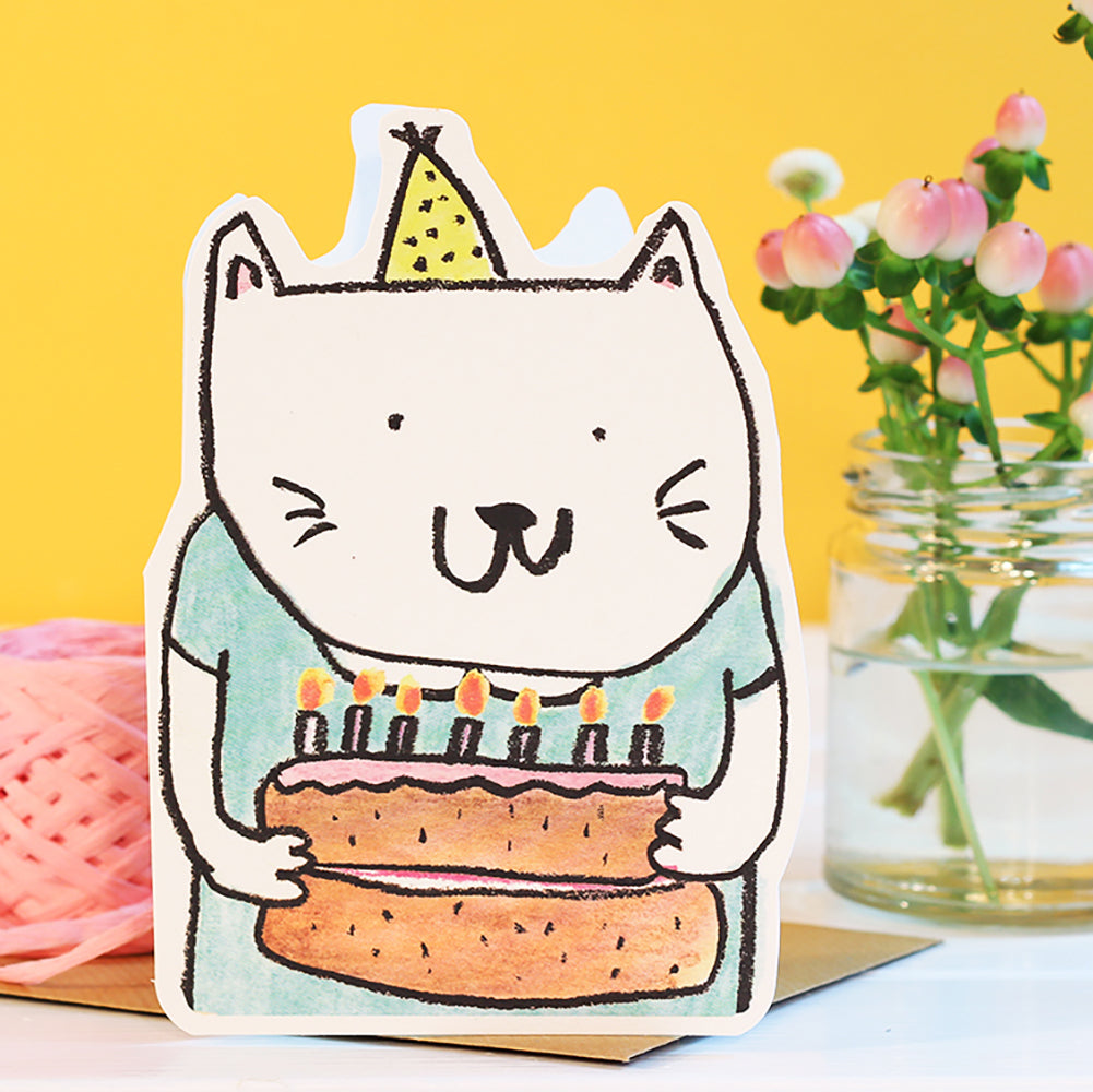 Card with a cat holding a birthday cake and candles against a yellow background. Handmade in the UK by Laura Skillbeck