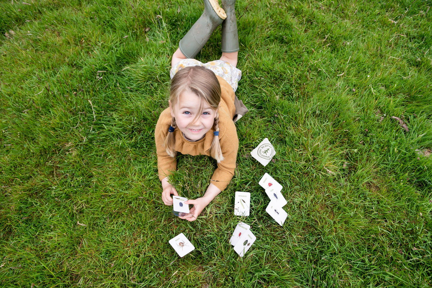 A child is laying on the grass, inspecting the illustrated British bug-spotter cards, packed with fascinating facts, and a pocket-sized guide to help identify your finds and understand their vital role in our ecosystem