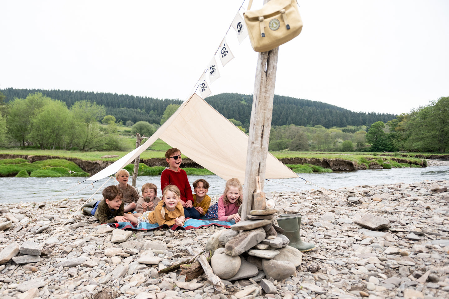 Pirate Den Kit including yellow cotton haversack, cotton calico tarpaulin, waterproof groundsheet, handmade wooden mallet, jute rope, steel tent pegs, pirate eye patch, and Jolly Roger bunting.