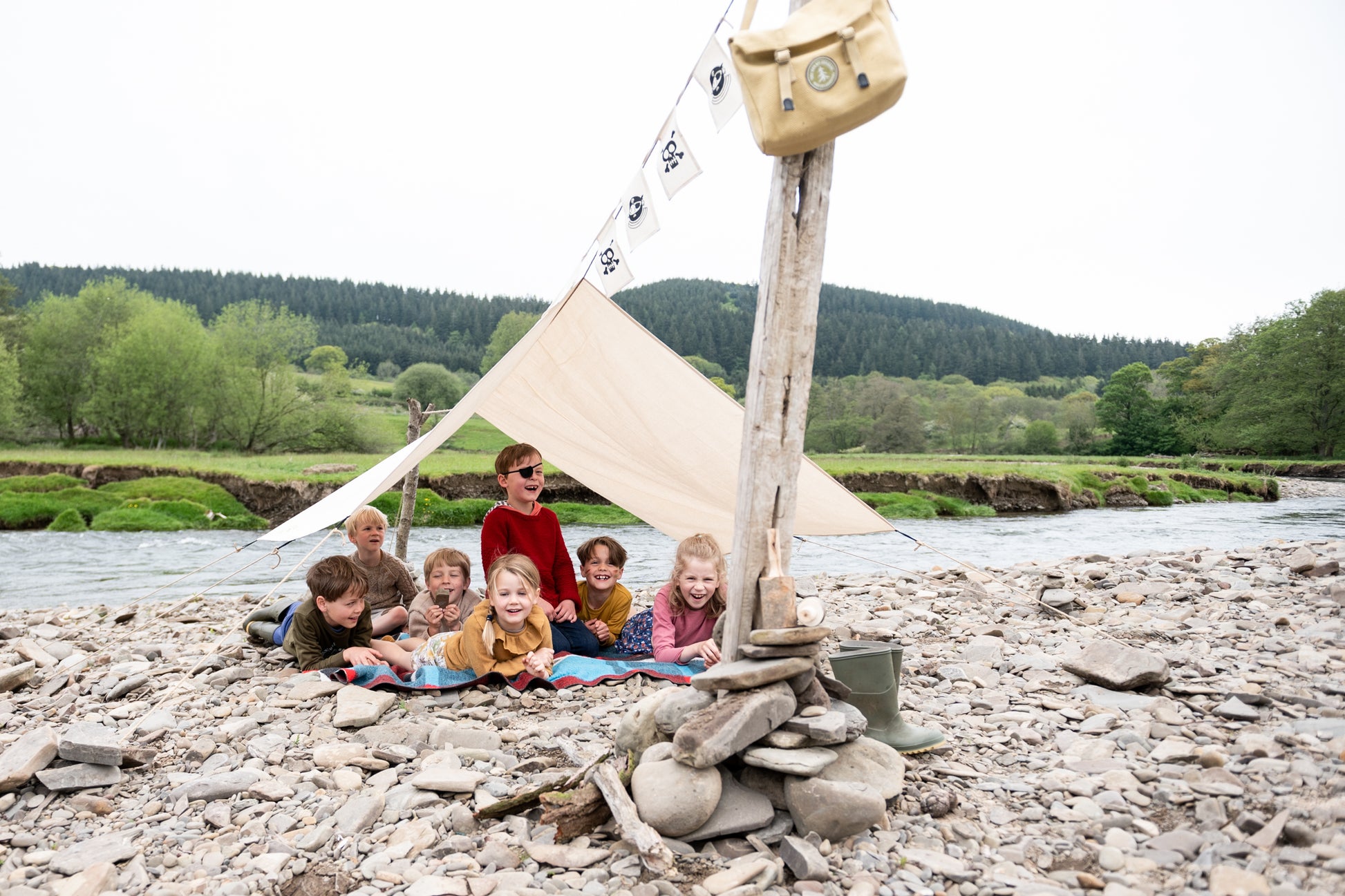 Pirate Den Kit including yellow cotton haversack, cotton calico tarpaulin, waterproof groundsheet, handmade wooden mallet, jute rope, steel tent pegs, pirate eye patch, and Jolly Roger bunting.