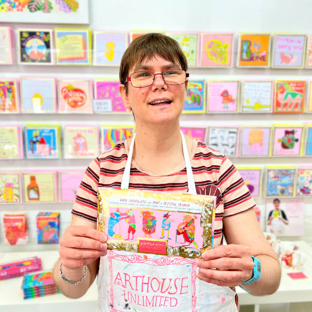 Person holding a chocolate bar with 'Arthouse Unlimited' branding in front of a display of greeting cards.