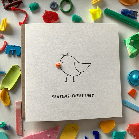 A square greeting card with a hand-drawn design of a bird on the front, titled 'Seasons Tweetings', placed on a background of colorful plastic pieces.