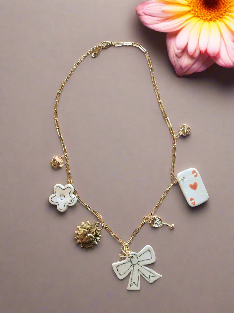 Celebrate Life Chunky Charm Necklace. Lovingly handmade, these ceramic charms are individually cut out from clay slabs and then hand-painted. They are hung on an 18K gold-plated chain.
Featuring a white and black bow, a white daisy, an Ace Playing card, gold wine glass, heart, eye and clover