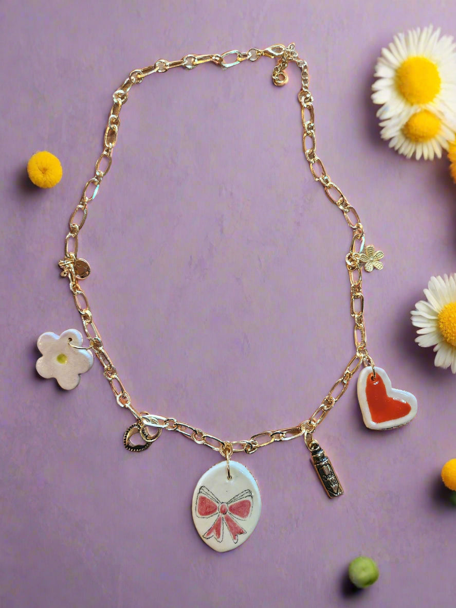 Celebrate Life Chunky Charm Necklace. Lovingly handmade, these ceramic charms are individually cut out from clay slabs and then hand-painted. They are hung on an 18K gold-plated chain. Ceramic heart, flower and bow. Gold charms flower, smiley face, heart padlock and "Drink me" bottle