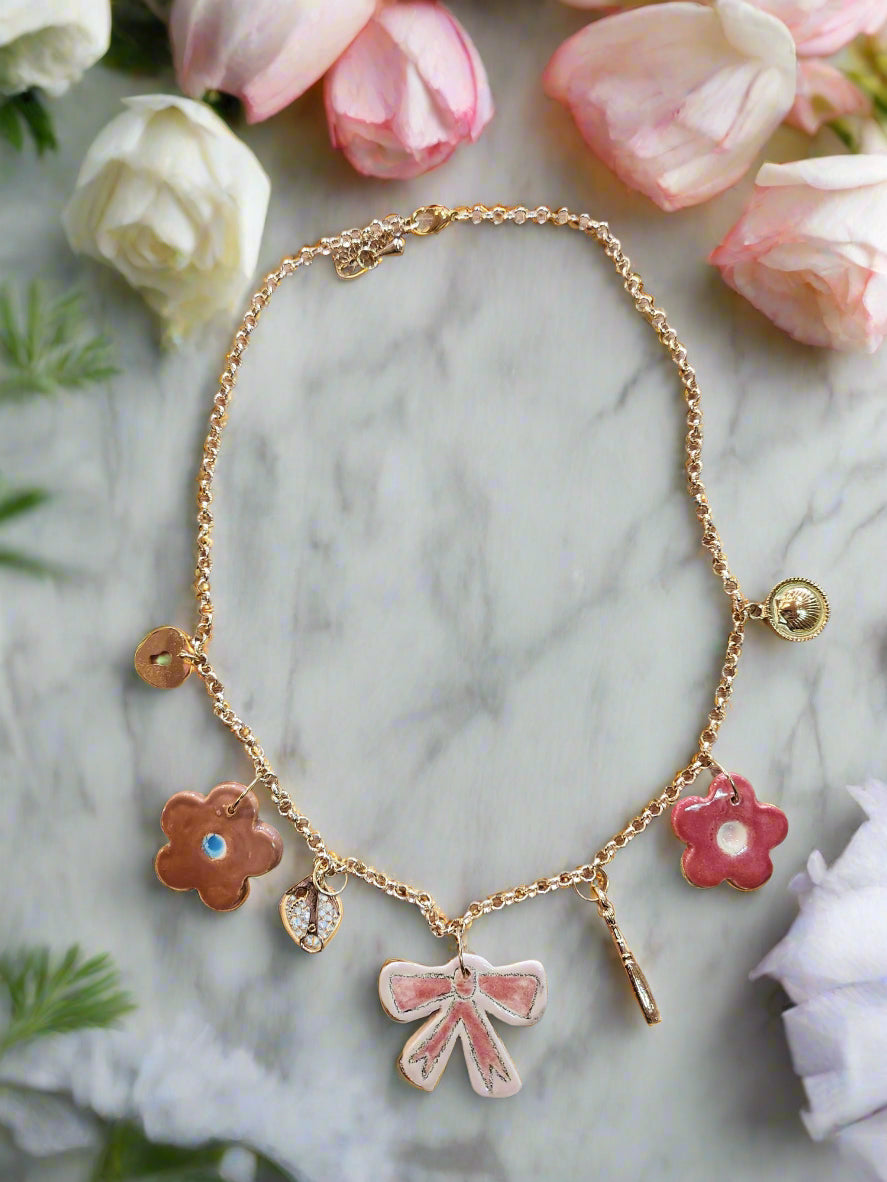 Celebrate Life Chunky Charm Necklace. Lovingly handmade, these ceramic charms are individually cut out from clay slabs and then hand-painted. They are hung on an 18K gold-plated chain. Ceramic Pink and brown flowers, pink ceramic bow, gold charms wine bottle, corkscrew, shell and heart padlock