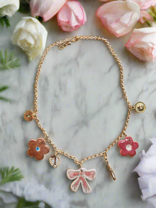 Handmade ceramic charm necklace featuring pink/brown flowers, pink bow, gold wine bottle, corkscrew, shell, and heart padlock charms on an 18K gold-plated chain.