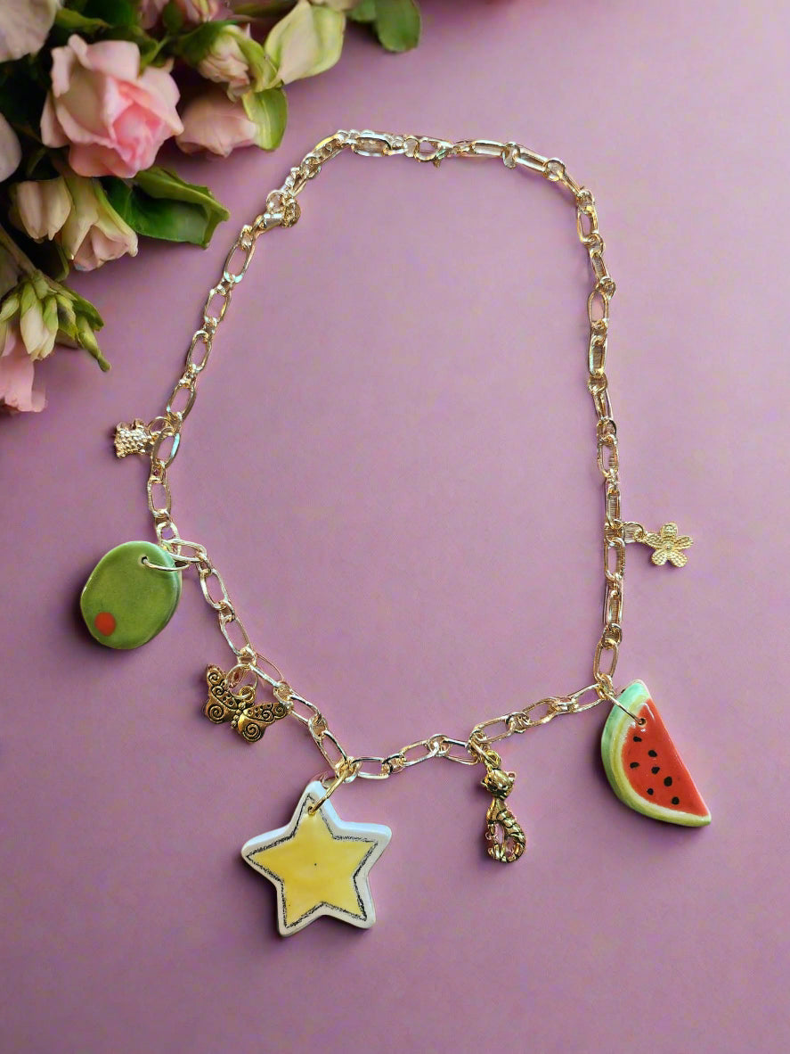 Celebrate Life Chunky Charm Necklace. Lovingly handmade, these ceramic charms are individually cut out from clay slabs and then hand-painted. They are hung on an 18K gold-plated chain. Ceramic yellow star, slice of watermelon and green olive. Gold chams, a flower, a cat, turtle and butterfly