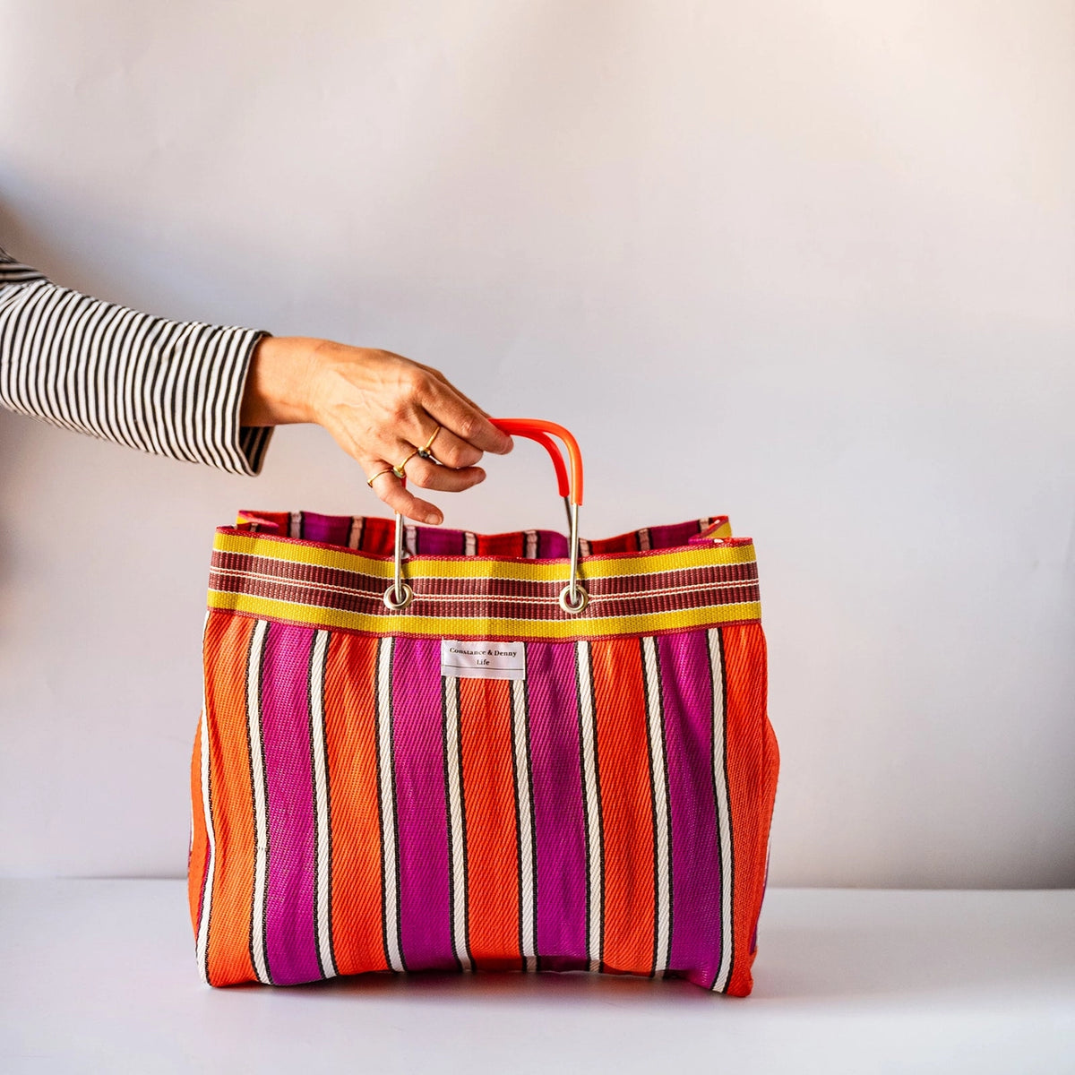 A colorful striped tote bag with vertical stripes in orange, magenta, yellow, and white, featuring a bright orange-red top handle, held by a person's hand against a plain light background.