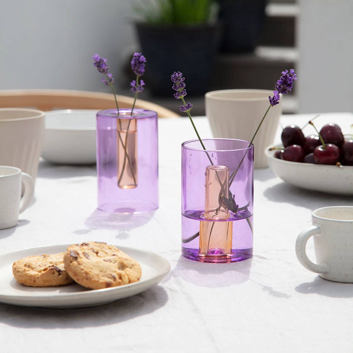 Two purple glass vases with lavender stems on a table with cookies and grapes.