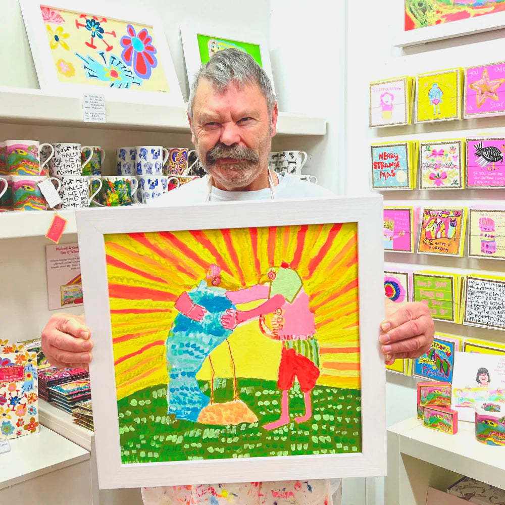 Man holding a framed colorful artwork in a store with greeting cards on shelves.