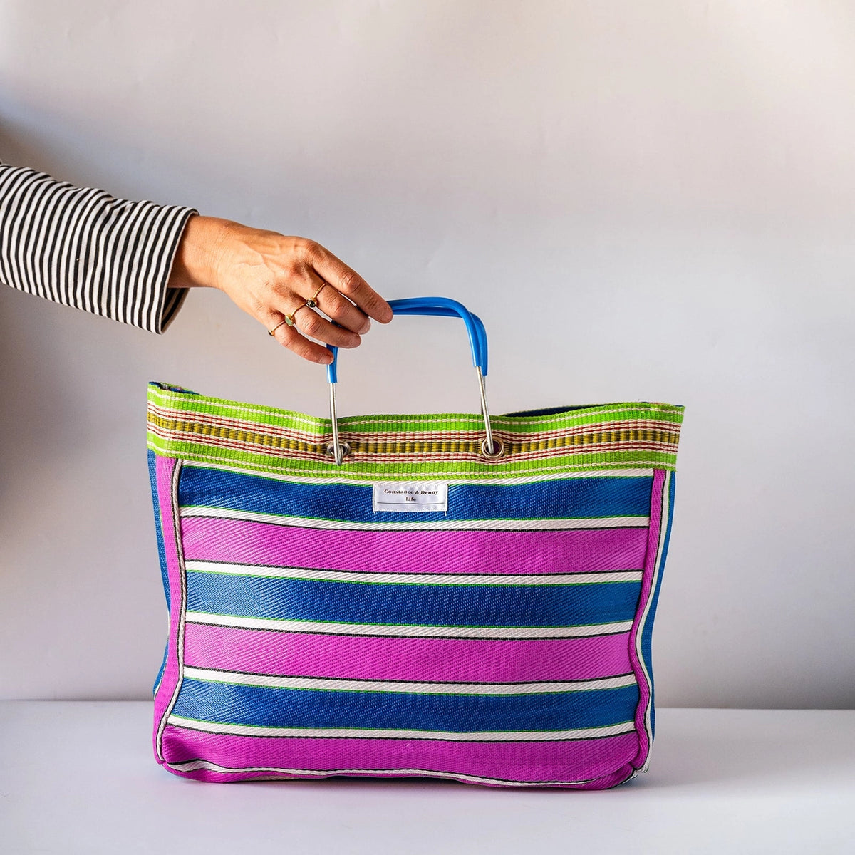 A striped tote bag with vertical bands of magenta pink, navy blue, and white, featuring a green and beige patterned woven border and a metal handle.