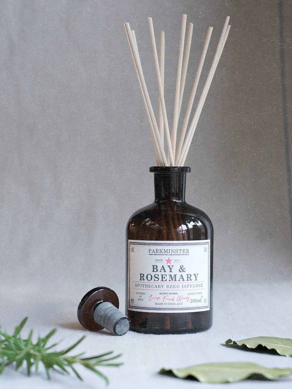 A  Bay & Rosemary reed diffuser bottle made of dark amber glass, with natural reed sticks inserted and a separate stopper lying on its side. The bottle is styled with rosemary and bay leaves on a light surface against a textured gray background.