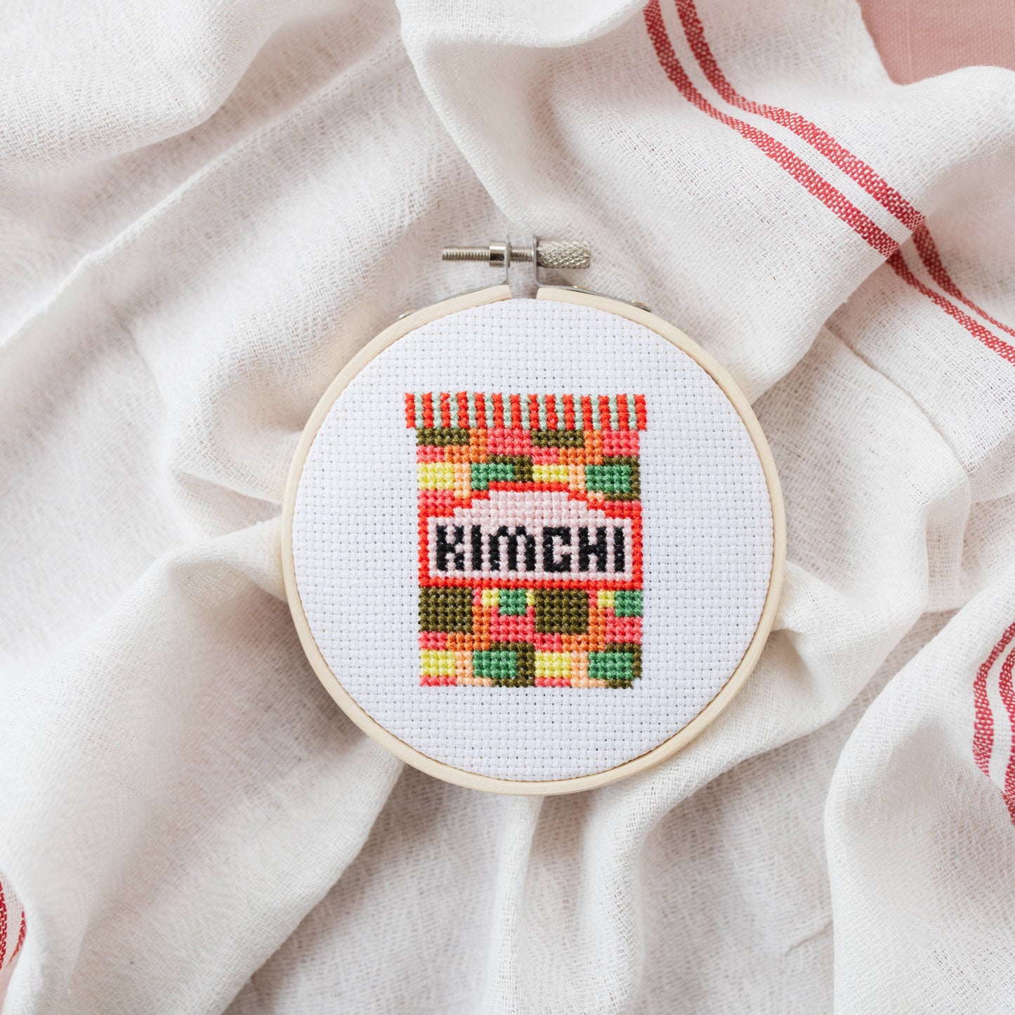 Finished Kimchi cross stitch art in its 11cm wooden hoop, displayed on a kitchen shelf as quirky decor.