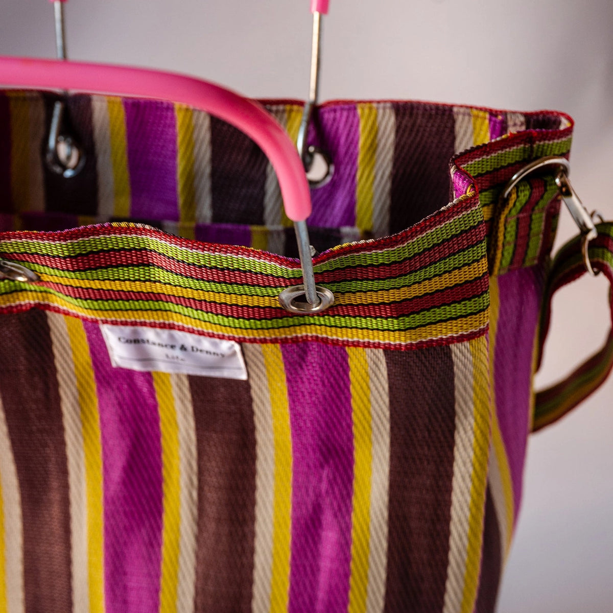 Close-up of a striped tote bag with vertical bands of purple, brown, yellow, green, and cream, featuring a pink plastic handle and metal grommets.
