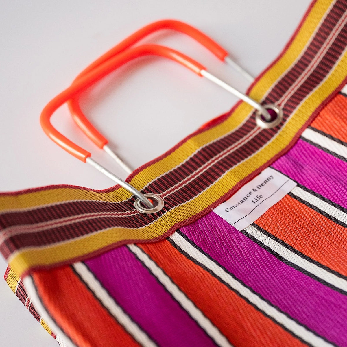 Close-up of a striped tote bag with bright orange handles, featuring bold stripes in orange, magenta, yellow, brown, and white. By Constance & Denny.