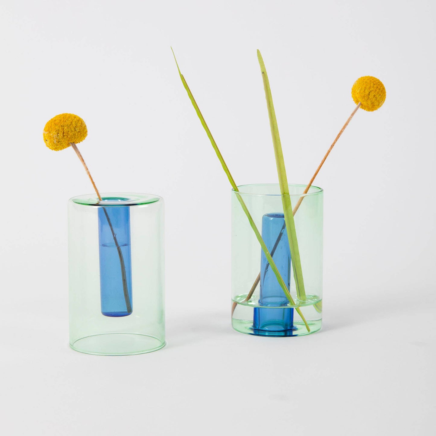 Two glass vases with blue centers and green exteriors, each containing a single stem of grass and a yellow flower on a white background.