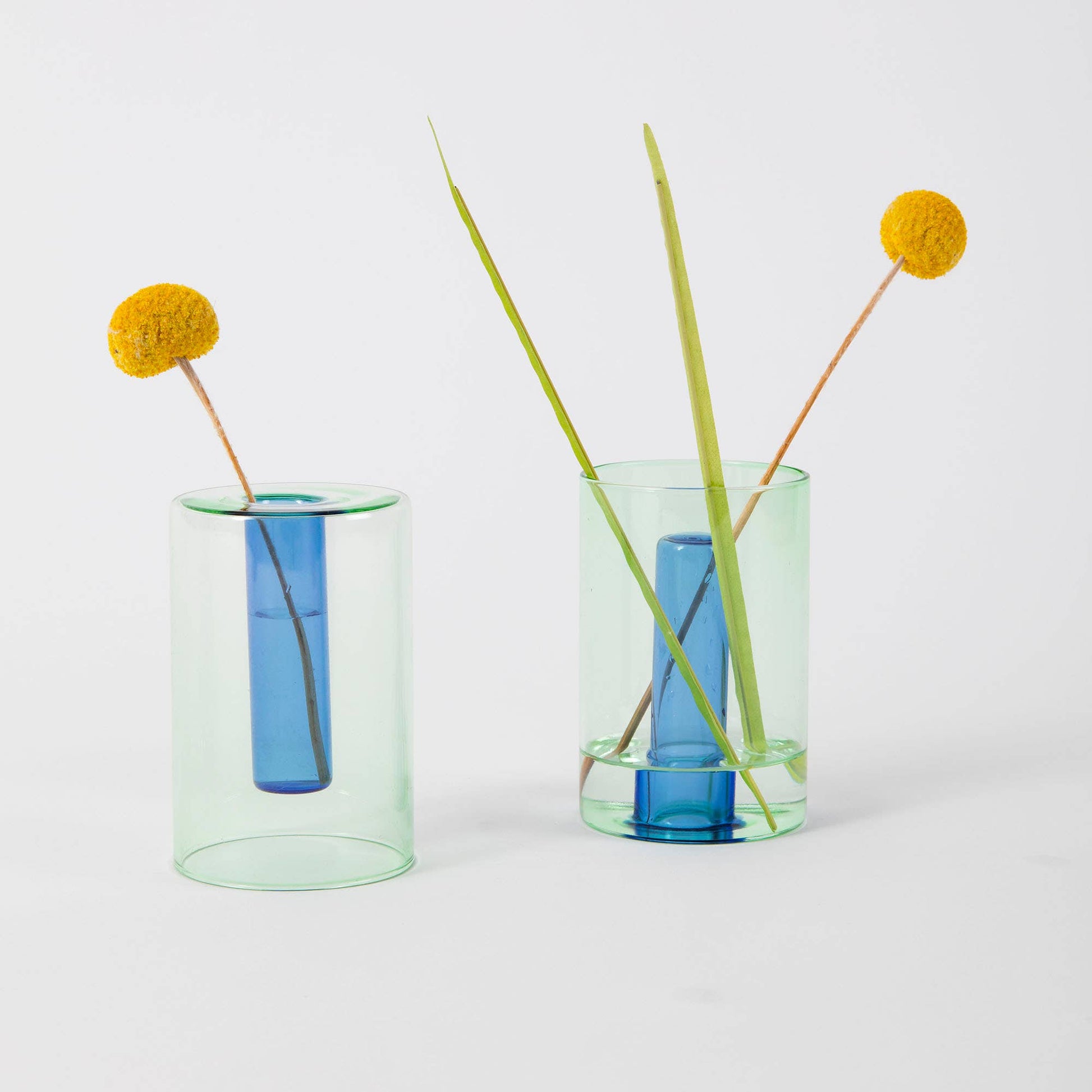 Two glass vases with blue centers and green exteriors, each containing a single stem of grass and a yellow flower on a white background.