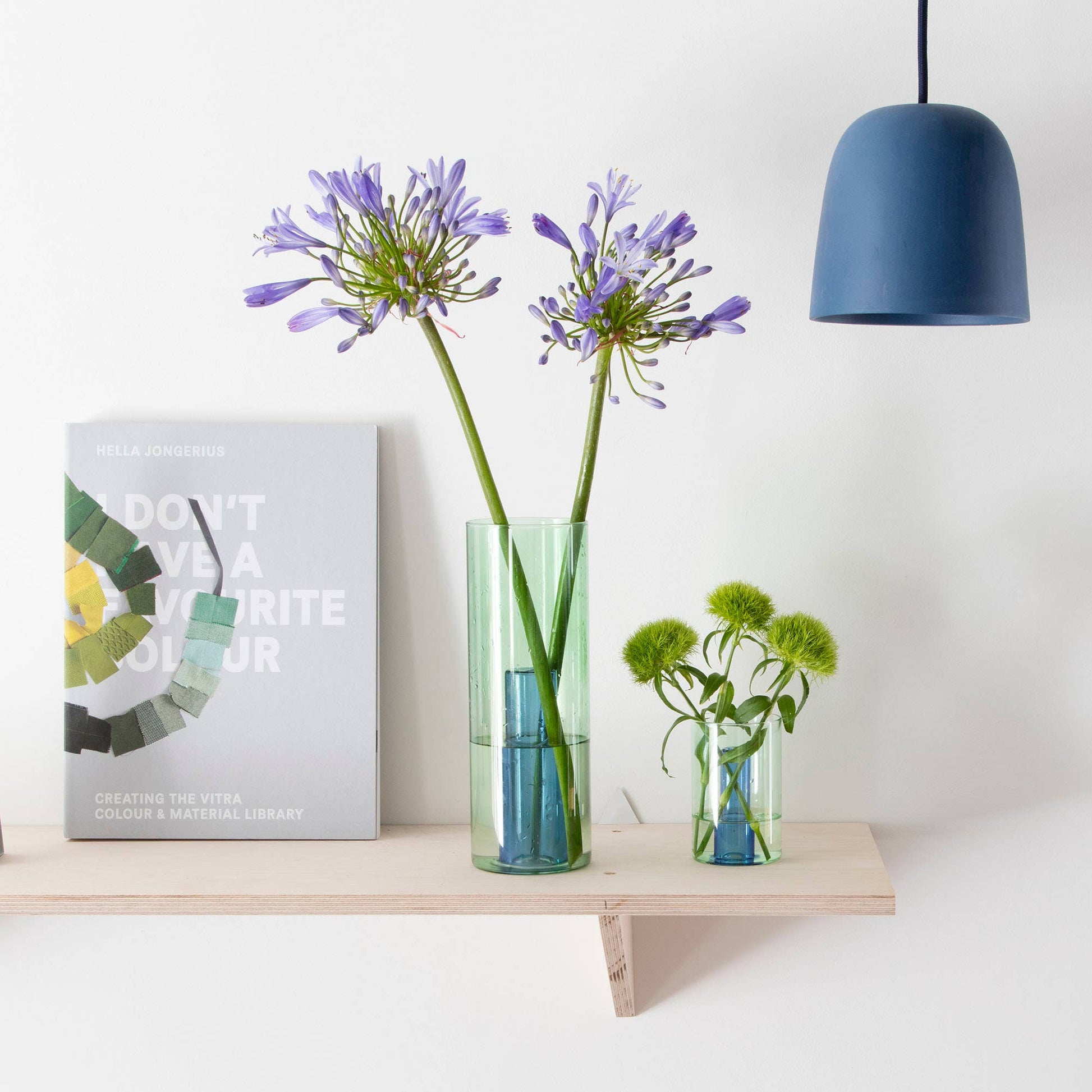Vases with flowers on a shelf against a white wall with a blue pendant light.