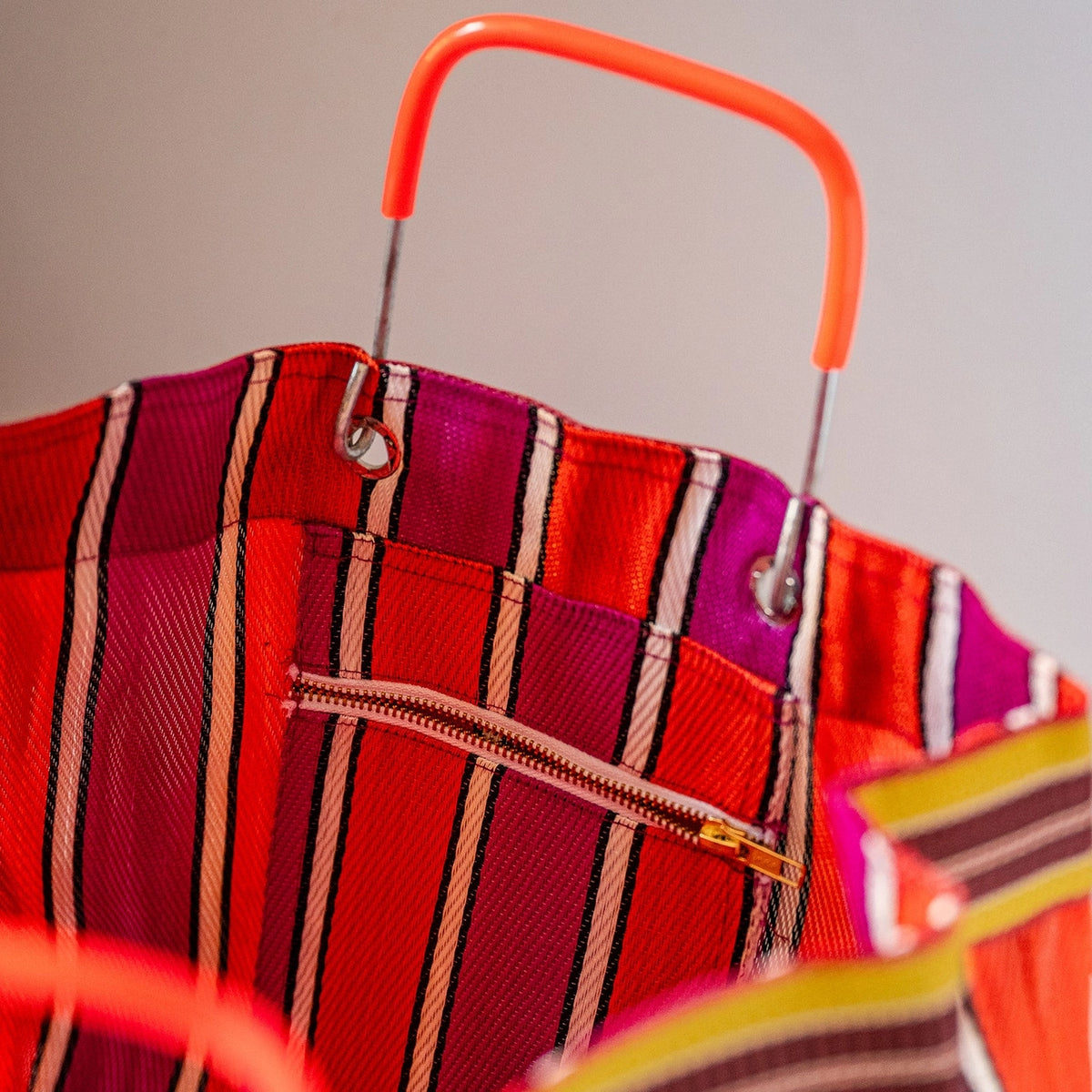 Close-up of a striped tote bag with a bright orange handle, featuring red, pink, and white vertical stripes and a zippered pocket.