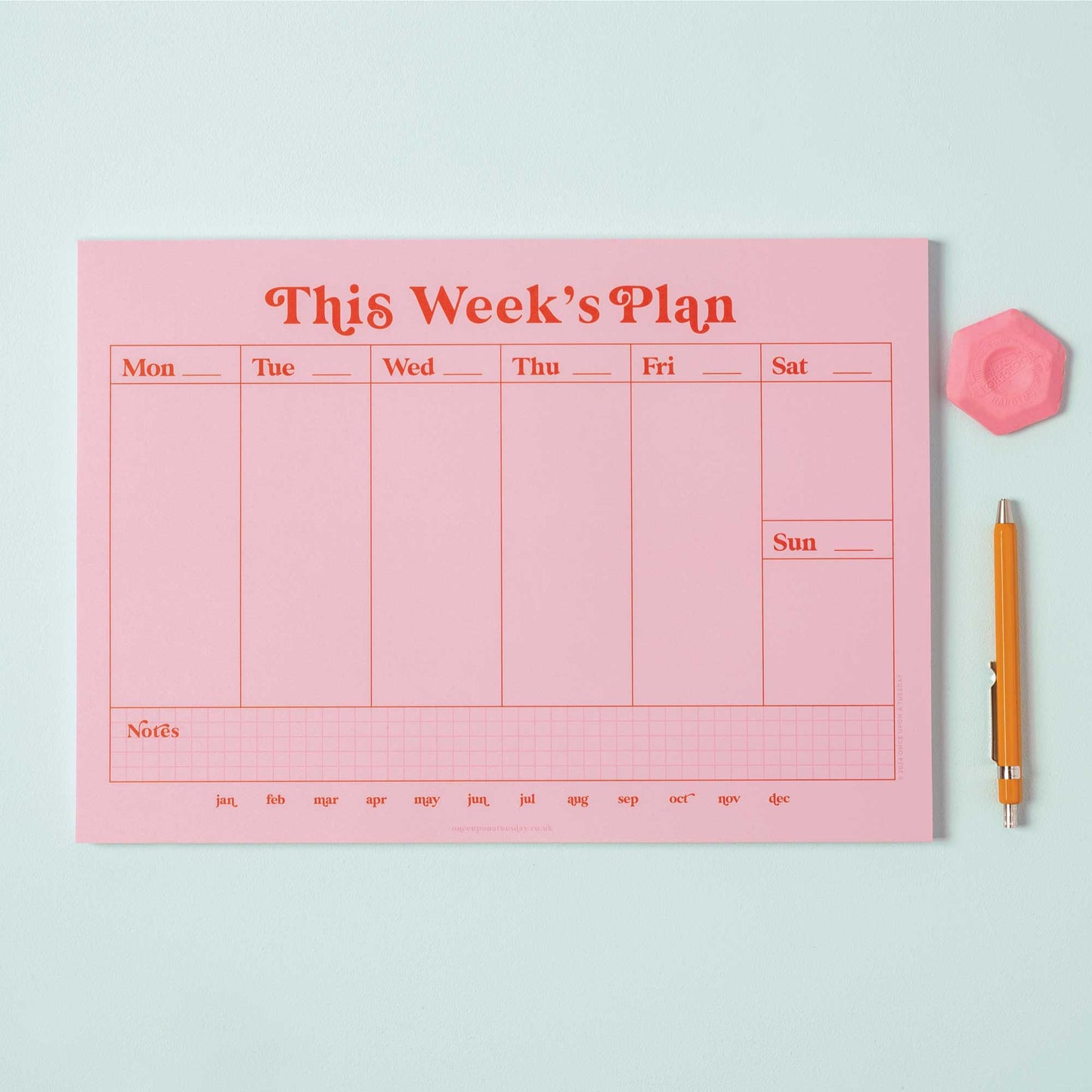 A4 Pink & Red Weekly Planner Pad on a desk, open to a blank week, with a pen, styled with a plant.