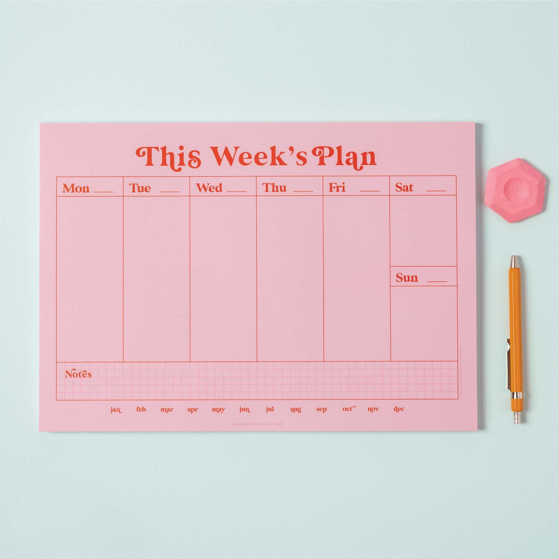 A4 Pink & Red Weekly Planner Pad on a desk, open to a blank week, with a pen, styled with a plant.