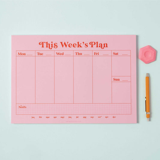 A4 Pink & Red Weekly Planner Pad on a desk, open to a blank week, with a pen, styled with a plant.