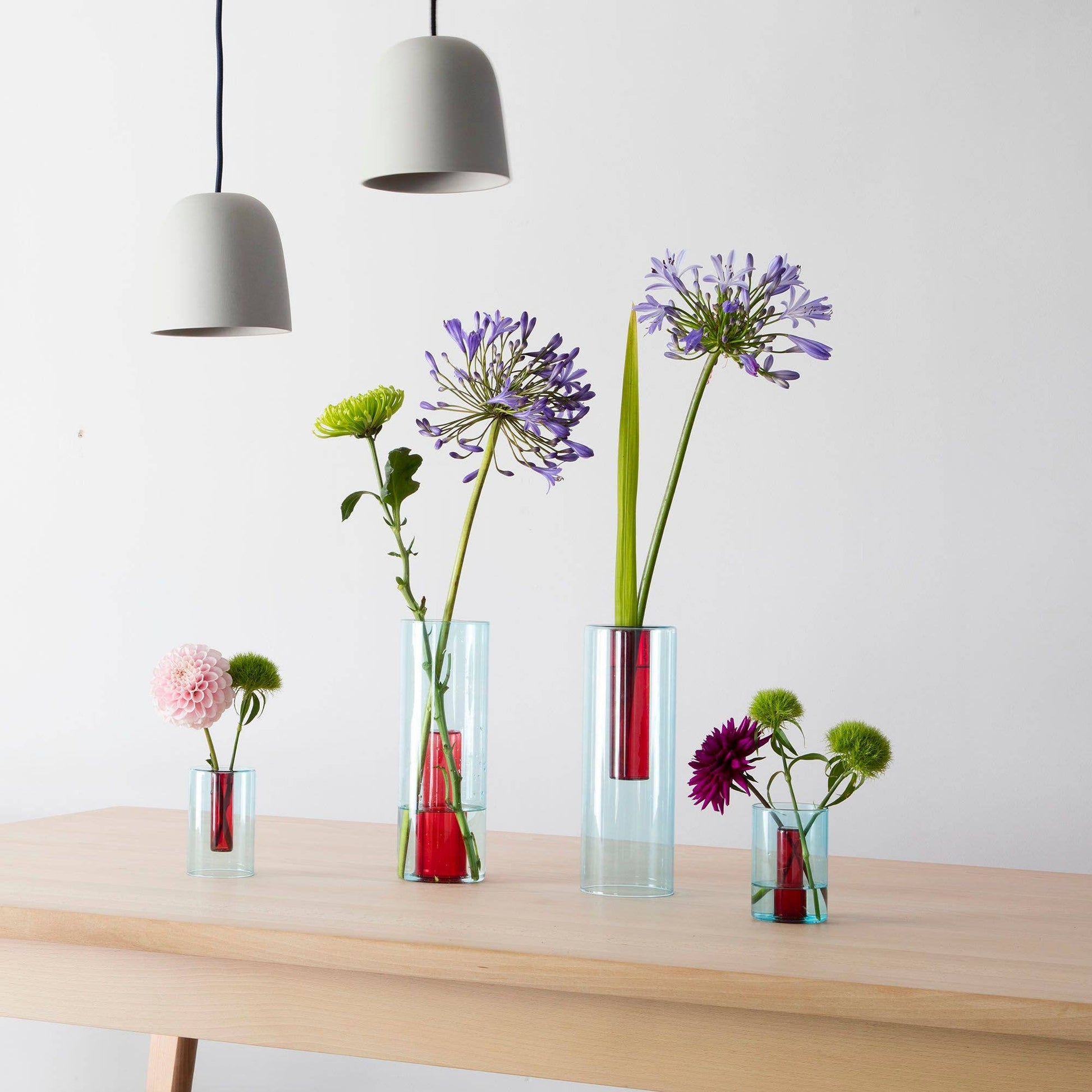 Set of glass vases with flowers on a wooden table against a white wall.