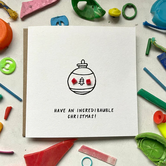 A greeting card with a hand-drawn and stamped design featuring the words 'Have an incredi-bauble Christmas!' and a Christmas tree ornament, accompanied by assorted colorful plastic pieces and a brown recycled envelope.