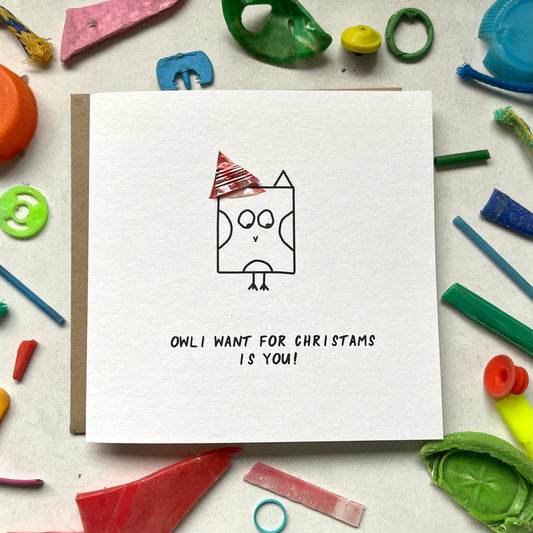 Handmade card with a drawing of an owl wearing a Santa hat, titled 'Owl I want for Christmas is you!' on a background of various colorful plastic pieces.