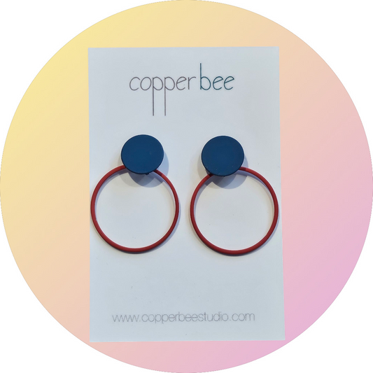 Close-up of marine blue and coral circle dot stud earrings on a 'Copper Bee' branded card with pastel background.