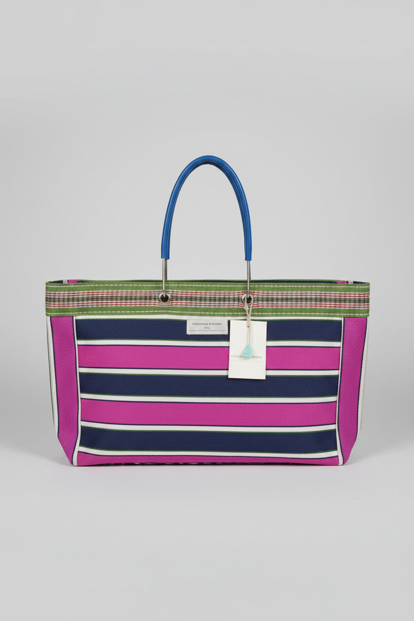 A striped tote bag with vertical bands of magenta pink, navy blue, and white, featuring a green and beige patterned woven border and a metal strap