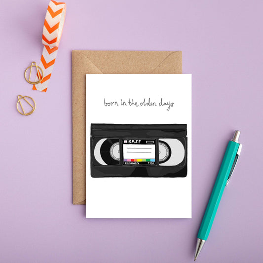 A birthday card with a VHS tape design, including a message that reads 'Born in the olden days', presented on a desk with a kraft envelope and a pen. Illustrated and printed in the UK by You've got Pen on your Face.