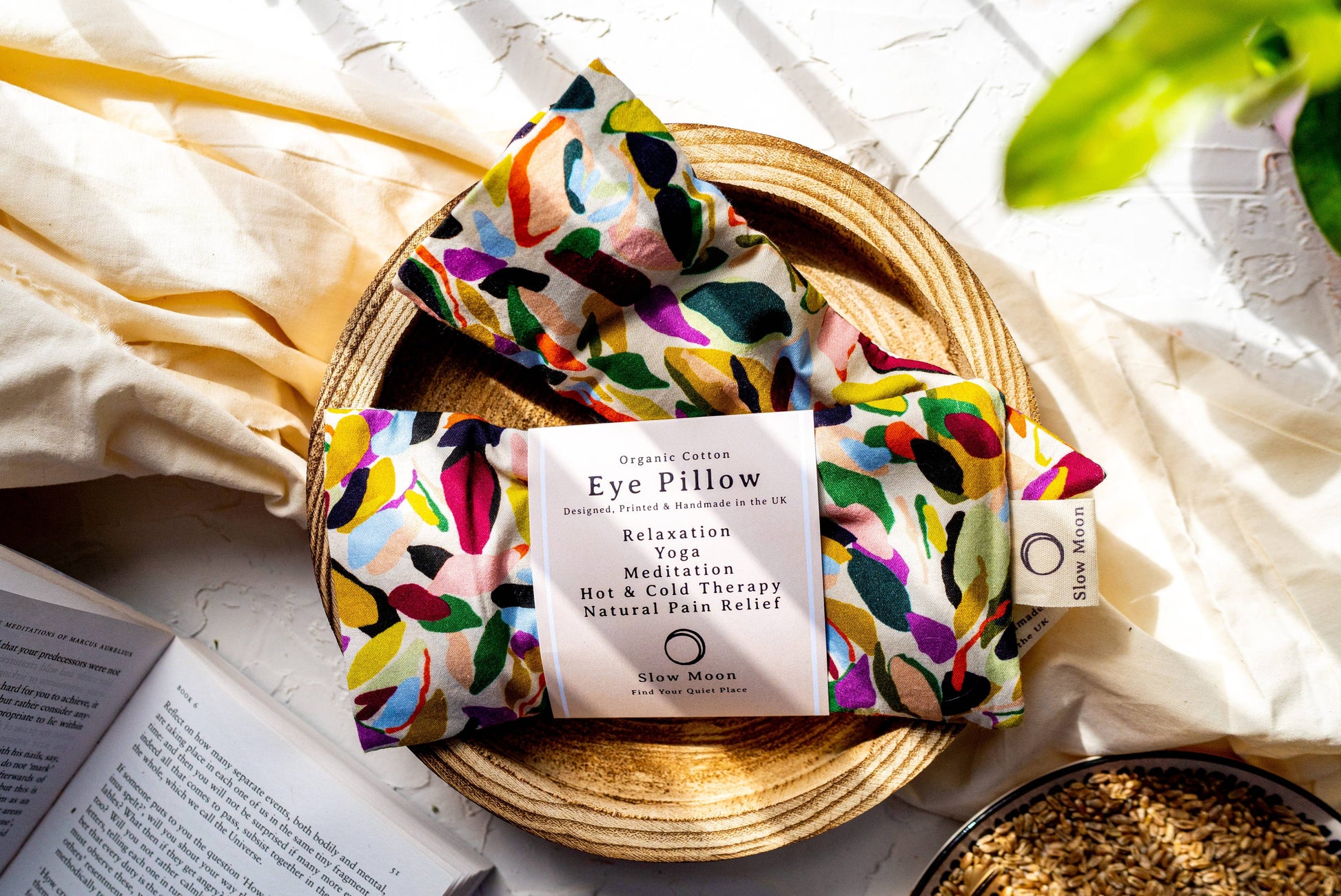 A multicolored organic cotton eye pillow with a floral pattern, displayed on a woven basket alongside a book and a plant.
